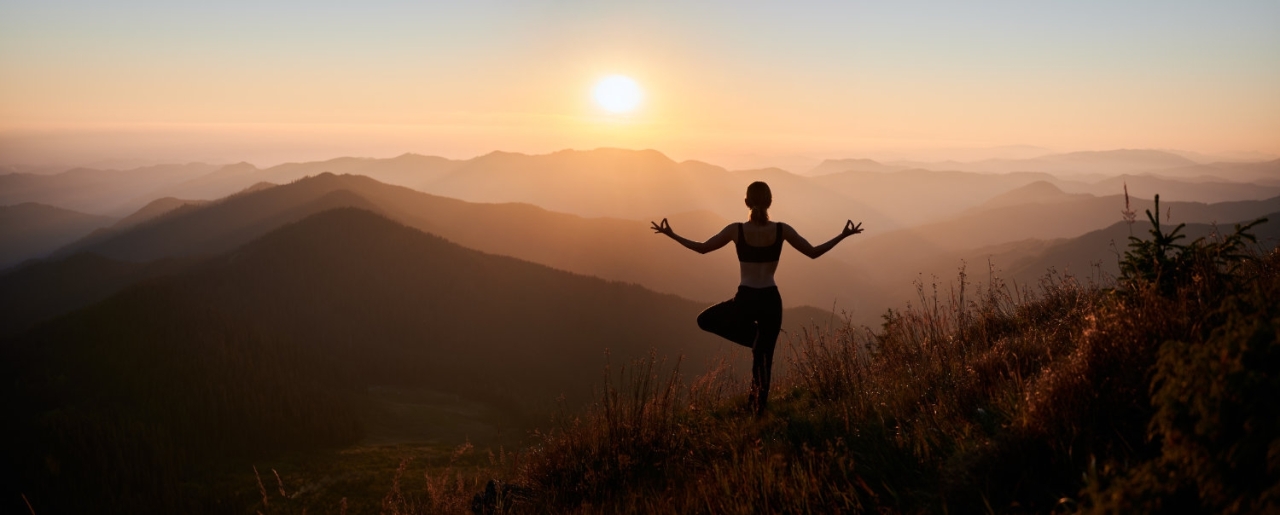 Woman meditating on nature with beautiful view. - Panoramic view. Back view of slim woman in black sportswear doing yoga exercise on one leg in mountains. Concept of yoga time at sunset.